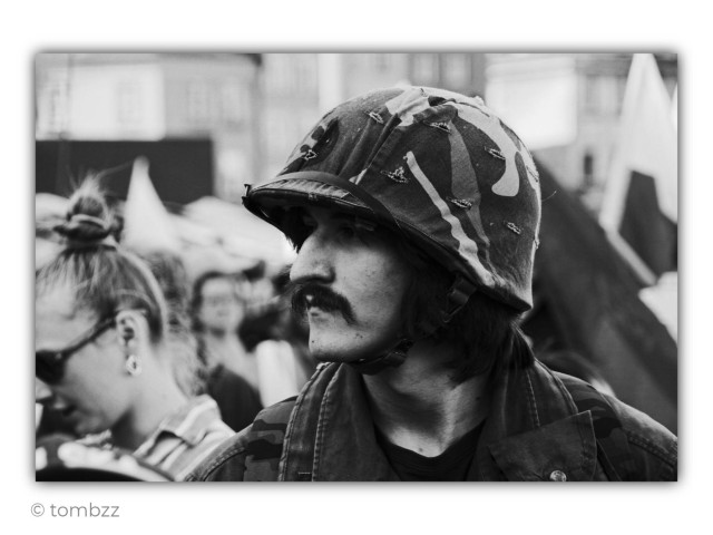 Black-and-white street photograph with a raw, documentary feel. In the center of the frame stands a young man with long dark hair and a thick mustache. He wears a military-style helmet covered with patterned fabric and a camouflage jacket. His profile is turned slightly to the right, his gaze sharp, focused, and filled with determination. His expression is serious, almost emotionless. The image evokes the visual language of Latin American revolutionaries, especially the iconic imagery associated with Cuban guerrillas. There is a subtle resemblance to Che Guevara, though presented here in a more modern and understated way.
The background remains softly blurred. A crowd of people can be seen along with fragments of flags or banners and hints of city architecture. On the left side of the frame, a woman wearing dark sunglasses looks downward, detached from the intensity of the main subject. The contrast between them adds tension to the scene. Strong contrasts and visible grain give the photograph the timeless atmosphere of classic black-and-white street photography.