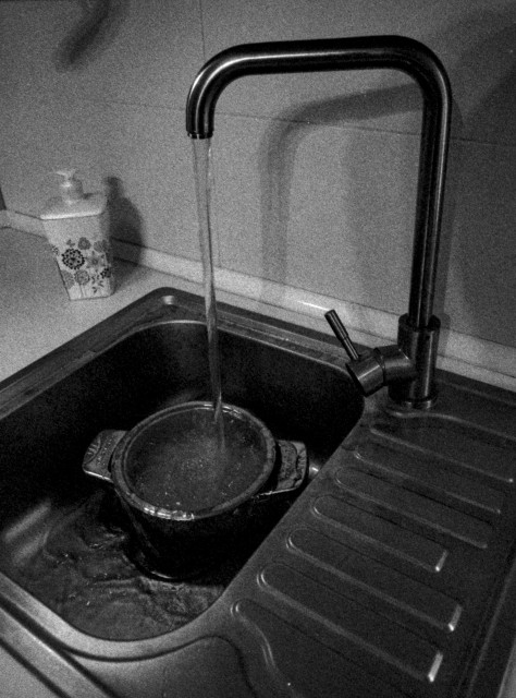 A grainy black-and-white photograph of a kitchen sink with water running from a modern faucet into a small pot placed below. The scene has a quiet, minimalist atmosphere, with soft shadows and reflections on the stainless steel surfaces. A soap dispenser sits in the background, while the textured film grain gives the image a raw, analog feel.