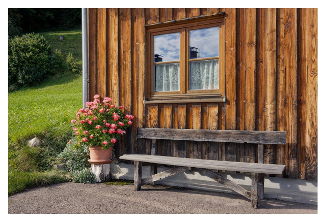 This is a sunny, peaceful outdoor photograph of a traditional wooden cabin. In the foreground, there is a gravel path. To the right, an old, weathered wooden bench with thick planks sits directly in front of the cabin wall. The bench looks sturdy but well-used. To the left of the bench stands a large terracotta flower pot placed on a tree stump. The pot is overflowing with bright pink and red geraniums in full bloom, creating a strong splash of vivid color. The cabin itself is made of vertical, warm brown wooden planks with visible wood grain and knots. In the center of the image is a double-pane wooden window with light-colored lace curtains visible inside. The glass reflects a bit of blue sky and clouds. Behind the cabin, a green grassy hillside slopes upward, dotted with small bushes and trees under bright daylight. The overall atmosphere is calm, idyllic, and picturesque — like a classic alpine or countryside scene on a beautiful summer day. The lighting is warm and natural, highlighting the rich textures of the weathered wood, the rough gravel, and the lush flowers.
