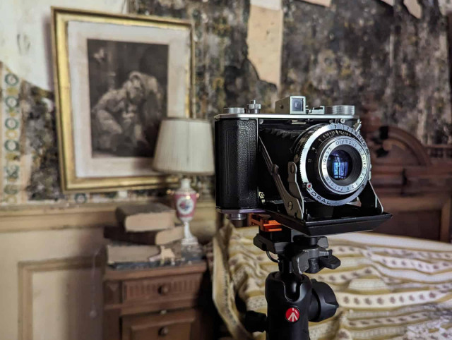 A vintage Olympus Six medium format camera on a tripod is ready to capture images in a room with peeling wallpaper and antique furniture.
