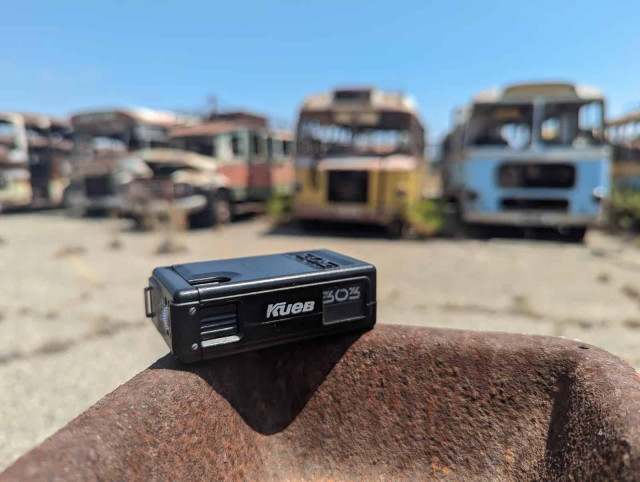 A black Kiev 303 subminiature film camera rests on a rusty surface, with blurred abandoned buses in the background under a blue sky