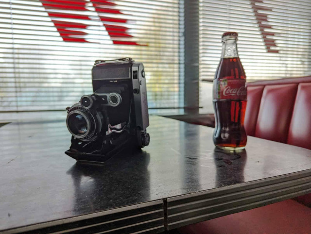 A vintage folding camera sits on a diner table next to a glass Coca-Cola bottle.