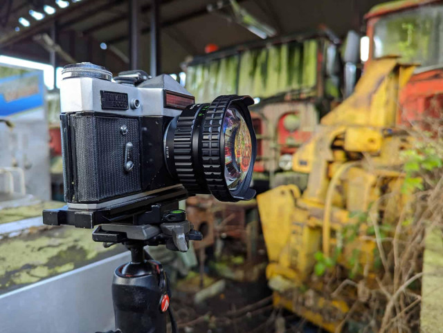 A vintage Zenith E film camera, mounted on a tripod, focuses on rusted, abandoned vehicles in a desolate yard.