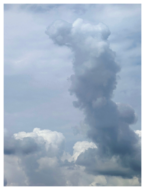 tall, vertical cumulus cloud rises against a blue-gray sky, with smaller fluffy white clouds at the base. The main cloud is dense and has a rounded top, resembling a column or plume, and is surrounded by faint, wispy clouds.