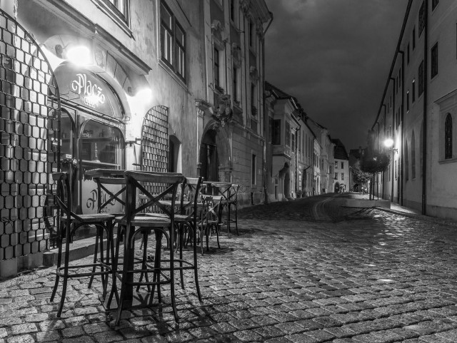 Nachts, s/w.
Nebenstraße mi Häuserzeilen und Kopfsteinpflaster. Vorn im Bild ein Tisch und typische Stühle eines Cafehauses, links davon Schriftzug am Haus.
Alles menschenleer.