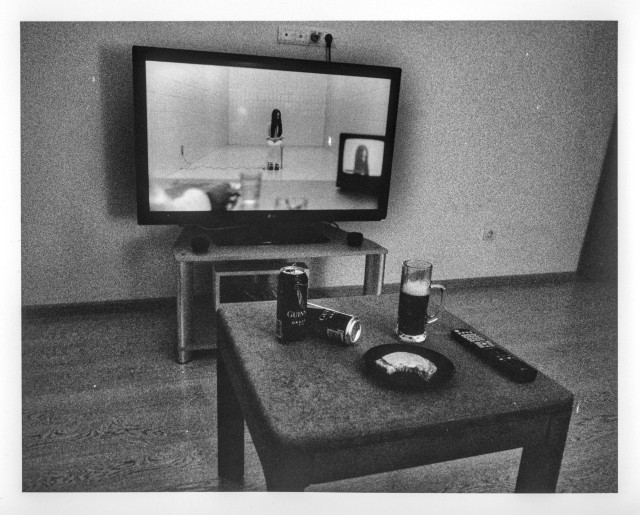 Black-and-white darkroom print showing a living room scene: a coffee table in the foreground holds two Guinness cans, a glass mug of beer, a plate with a sandwich, and a TV remote. In the background, an LG flatscreen TV displays a scene from the horror film "The Ring," with a small CRT television visible within the larger screen. Heavy grain characteristic of push-processed film.