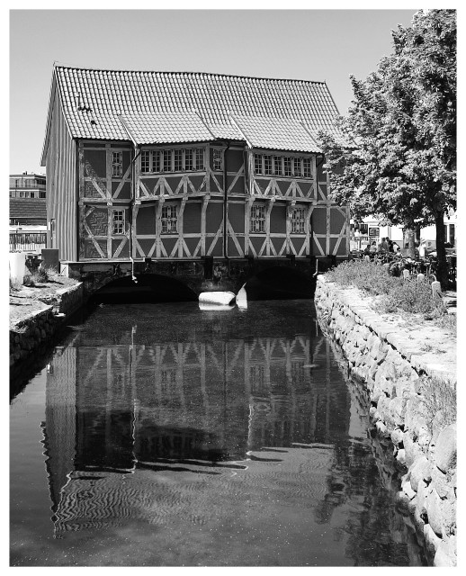 Schwarz-Weiß-Foto im Hochformat mit weißem Rand. Zu sehen ist ein sehr altes Fachwerkhaus ein einer kleinen Brücke, es spiegelt sich im Wasser. Das Haus ist schief, die Balken und Fenster ebenfalls. 