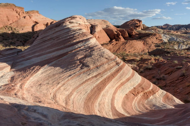 A color landscape photo of a sandstone formation. The eroded sandstone block in the foreground is shaped like a cone cut in half. It's narrow on the right and wide on the left. There's a knob on the far side at the top. There are many layers of brick red, beige, and tan sandstone curving horizontally across the cone like depression. In the background are large boulder like red sandstone formations under a blue sky with a few clouds.