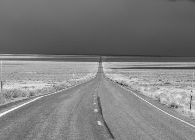 A black and white landscape photo looking down an empty two lane highway. The highway curves in from the right and is straight until it disappears over the horizon. Short dry grass grows on each side of the highway. It is sunny on the road in the fore- and middle-grounds and In the distance you can see finger like shadows of clouds crossing the road. The sky in the distance is uniformly dark with rain clouds. 