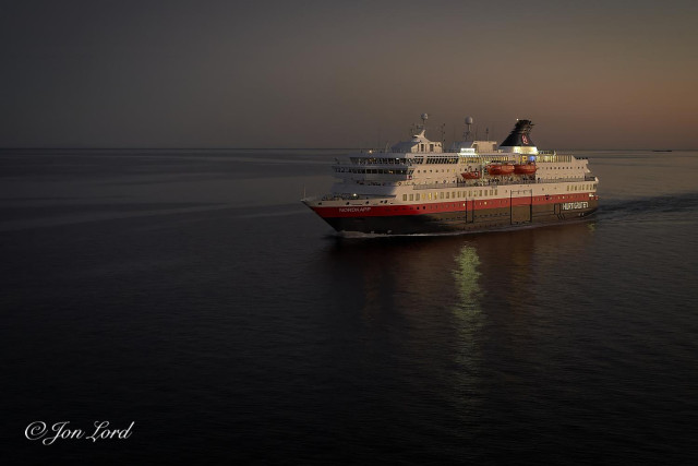 This is a colour photo in landscape format of a Norwegian coastal ferry sailing through a dead calm sea during the near midnight sun giving the appearance of a prolonged sunset before the dawn: the night never getting truly dark. Vestfjorden, Norway (2018).

The calm expanse of cobalt blue, almost black sea rises upwards to the three-quarters up level. Above a crisp horizon is a clear and cloudless sky, near dark in the upper left corner and while in the upper right corner the sun being below the horizon gives a transition of multiple gentle peach red colours, graduating to a dark blue at the top. Much of the sea's surface is mirror flat with a few small ripples in the lower half of the image caused by the lightest breath of wind. In the centre and to the right is an oblique view of a coastal ferry, underway from right to left. The bow and port (left) side is in view and with the ship occupying half the width of the photo. The vessel has a black hull with a thick, bright red stripe on top and with a white superstructure above. Hanging from davits are three bright orange lifeboats. Aft, on the upper deck is a raked back, black funnel with the shipping lines logo: a white 'H' against a black circular background and light by a spot light. On the bow set in the red strip is the ships name: 'NORDKAPP' in bold white lettering while at the stern is the shipping line's name 'HURTIGRUTEN' again in white letters. The sky is reflected on the white of the ship giving it a pink glow.