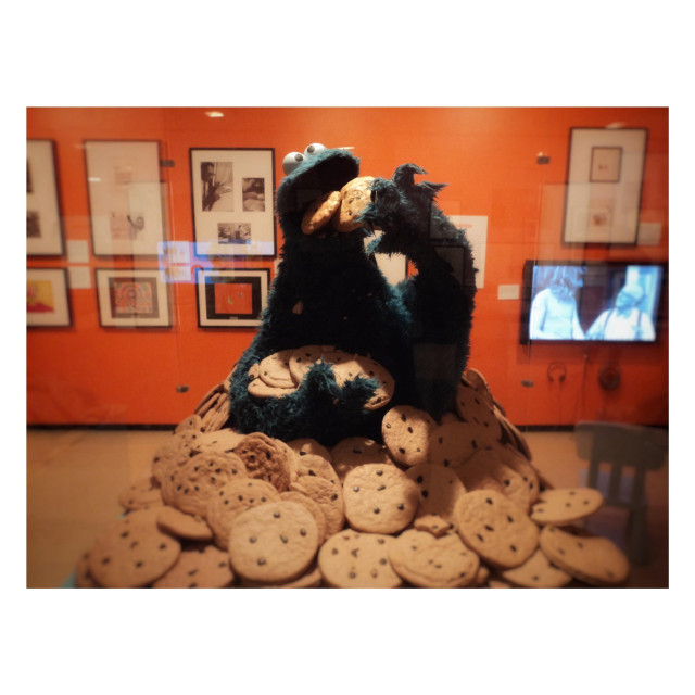 Photo of a display case featuring the furry blue muppet Cookie Monster from Sesame Street, half-buried in a pile of chocolate chip cookies and stuffing a couple of cookies into his open mouth. Behind is an orange wall displaying some framed photos and a TV screen showing a black and white scene from the show. Taken at an exhibition on Sesame Street at the New York Library for the Performing Arts many years ago.