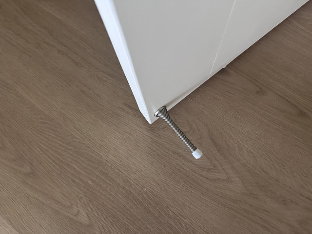 A spring-style doorstop ("thwonger") attached to a white door, against a wood floor.