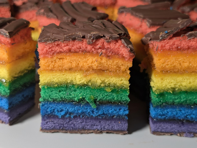 Lots of cake-slice-like cookies stand on a platter before the camera. They're rainbows, with top-to-bottom red, orange, yellow, green, blue, and purple layers, all vibrant! They're sandwiched with apricot jam, and bordered top and bottom with a chocolate layer.