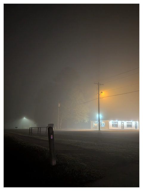 foggy night scene with a faintly lit street, glowing streetlights, power lines, and a brightly illuminated building in the background. a wooden post and a fence in the foreground, with trees partially obscured by mist.