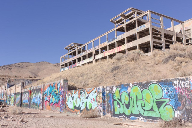 A color landscape photo of a very large concrete structure that is little more than the skeleton of a large ore processing facility. The structure sits high on a hill, is long, and is three stories high. There are two turret like additions on the top, one on the far left and one on the far right. The camera is looking at the the corner of the structure which is on the right side of the frame. The hillside is covered in dry grass. At the base of the hill, perhaps 60 feet below the structure is a long concrete wall covered in graffiti. The sky is clear and deep blue.