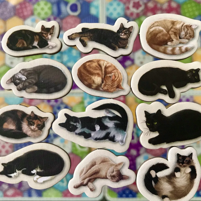 Cat tokens for the Calico board game. 12 cardboard tokens, each with a different cat posing, with a white border around them. The background is a colourful board in the style of a calico quilt.
