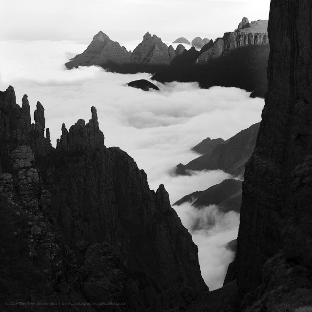Dark, needle-like basalt pinnacles rise from the foreground escarpment edge in sharp silhouette, framing a view across a vast sea of cloud that fills the valleys between multiple receding mountain ridges and serrated peaks stretching to the horizon. The black and white treatment deepens the contrast between the near-black foreground spires and the luminous white ocean of cloud below, with successive ridgelines fading progressively lighter into the pale background sky.