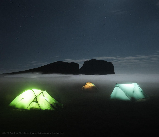 A night photograph of three illuminated tents glowing in green, amber, and blue-white on a high mountain plateau, with a layer of mist partially obscuring the base of a dark, flat-topped rock formation silhouetted against a star-scattered sky. The tents sit half-engulfed in the drifting cloud layer, their warm inner light diffusing softly into the surrounding mist. The combination of the glowing camp, the floating rock buttress, and the star-filled sky above creates a magical, otherworldly atmosphere on the Drakensberg plateau.