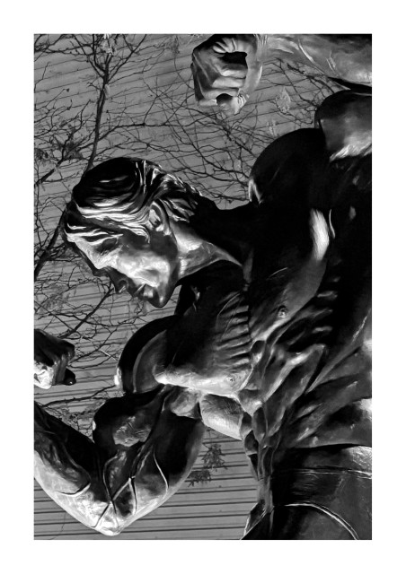 Black and white photo of a statue of Arnold Schwarzenegger at his peak bodybuilding physique, shot from the waist up. Arnold flexes his muscles in his signature “double biceps” pose, with waist twisted and both arms curled, one raised higher than the other. A spotlight brings out the contours of the statue in contrasting light and shadow. In the background are slender tree branches and a building facade with ribbed metal siding.