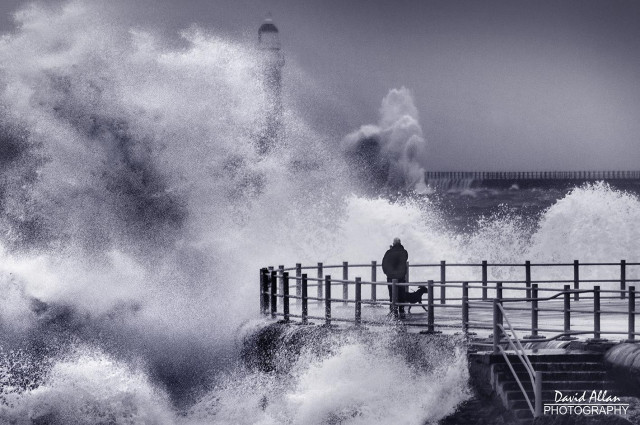 One man and his dog standing on the promenade at Seaburn, Sunderland in NE England. It's a June day, summer is almost here, but the sea conditions look anything but inviting. Waves crashing all around, leaden skies and a scene that looks more like a mid-winter storm. Certainly not a day for deck-chairs and picnics on the beach.