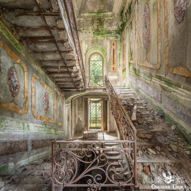 A crumbling, moss-covered grand staircase with ornate rusty railings descends in an abandoned Italian manor