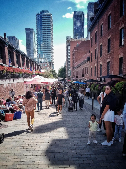 People are seen walking and sitting along a wide open walkway between buildings on a sunny spring day. Tall buildings can be seen in the distance. 