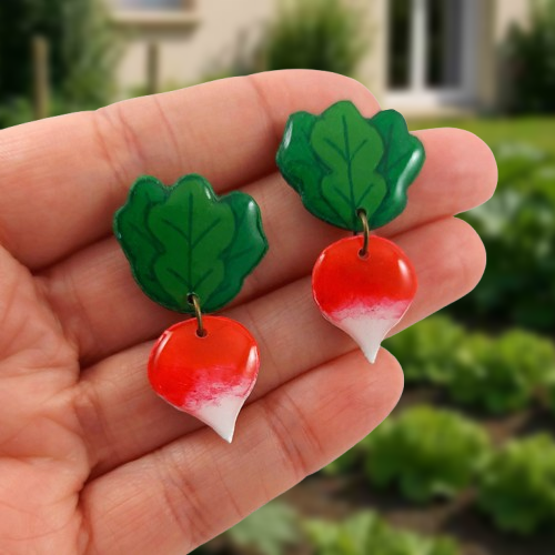Puces d'oreilles fantaisie pendantes en forme de radis ronds, et ses feuilles.
Les perles sont réalisées en CD recyclé, découpées et peintes à la main, puis recouvertes de résine.
Les tiges sont en acier inoxydables.
Ces puces d'oreilles sont tenues entre les doigts tendus d'une main ouverte. En arrière-plan, un potager devant une maison.
