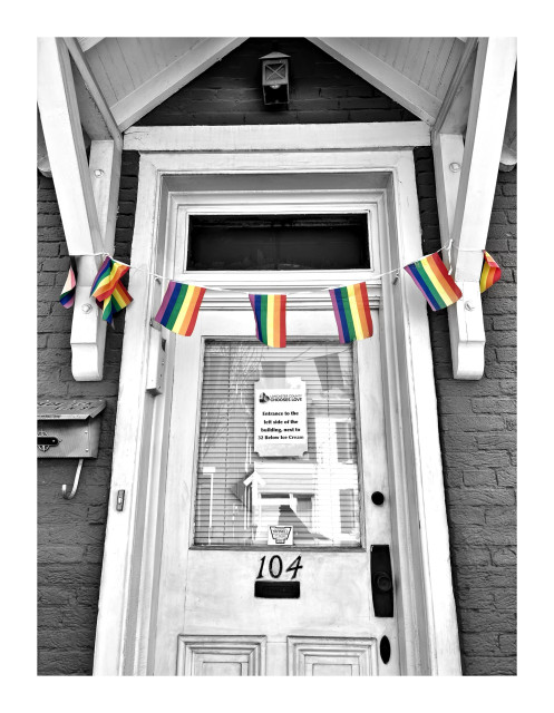 Photo of a paneled wooden front door in the brick facade of a house, flanked by the brackets supporting a wooden overhang. A little lantern is hung in the triangular space between the overhang and the door lintel. Strung across the doorway between the two brackets is a  length of string from which hang several miniature rainbow flags. Taped to the glass panel of the door is a sheet of paper that reads “Lancaster County Chooses Love” and some directions to use a different entrance to the building. The house number “104” is displayed below the glass. The photo is desaturated except for the rainbow flags, which pop out colorfully against the monochrome.