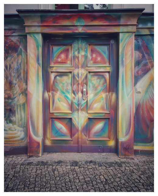 Farbfoto im Querformat. Zu sehen ist ein Teil einer Hauswand, der komplett mit einem Auftragsgraffiti bemalt ist. In der Mitte des Fotos sieht man eine ebenfalls bemalte Tür zum Souterrain, wie sie für Kreuzberger Altbauten typisch ist.