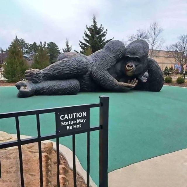 a giant statute of a gorilla on a minigolf course that is lying on its side like an odalisque with a sign on the gate to the course that says: caution - statue may be hot 