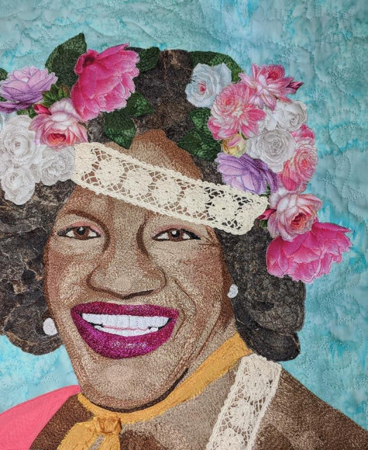 Pictorial quilt featuring a smiling image of Marsha P Johnson in a floral headpiece set against a light blue background. The quilt is heavily quilted using the thread painting technique to get a fairly naturalistic representation of Johnson while the flowers appear to be broderie perse appliqué and actual lace trim has been added for additional dimension