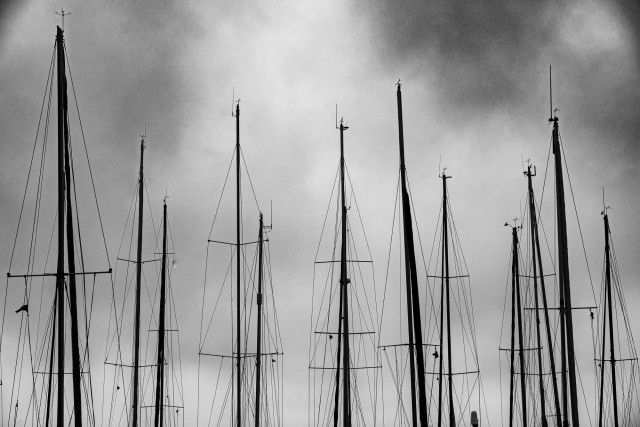 Zu sehen sind mehrere Masten von Segelbooten (oder sagt man Yachten?) - ohne Segel, nur mit Halteteauen - vor einem bewölkten Himmel. Das Bild ist SchwarzWeiß.