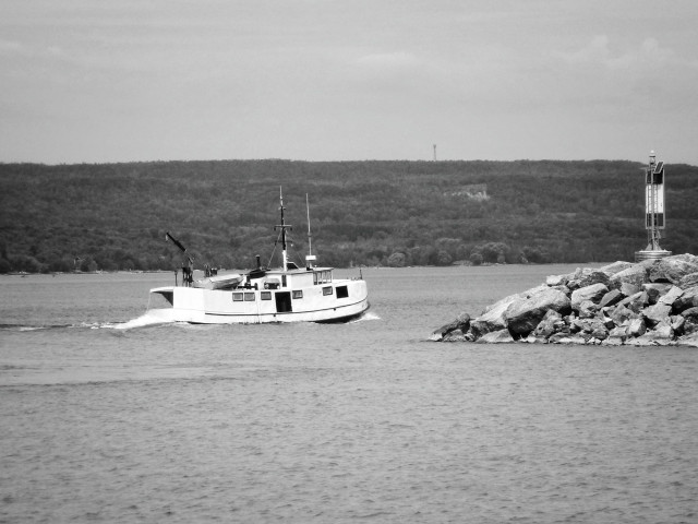A white fishing boat heads out onto the water of a large lake passing by a stone breaker wall with a navigation sign on the end of it. The hills of the tree covered shoreline can be seen in the background. This is a black and white image.