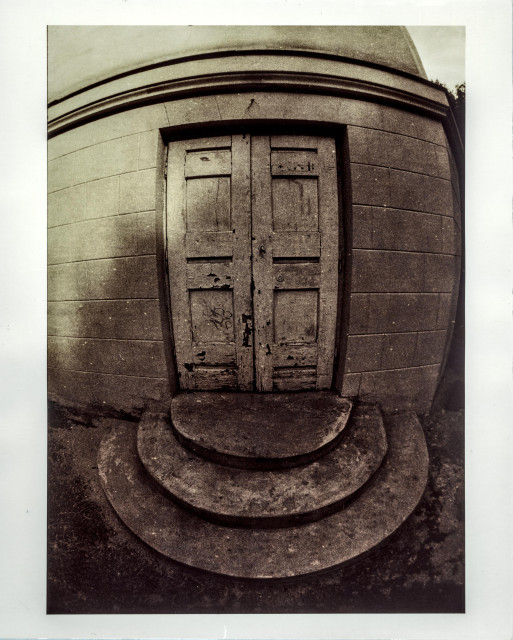 Lith print photograph of a weathered double wooden door set into a stone wall, shot with a fisheye lens. The extreme barrel distortion bends the horizontal lines of the stone masonry outward, giving the building a swollen, spherical appearance. The door shows heavy wear — peeling paint, scratches, and faded graffiti on the lower panels. Three semicircular stone steps lead up to the threshold. The warm sepia-brown tones typical of lith printing deepen toward the edges, with a pale border surrounding the print.
