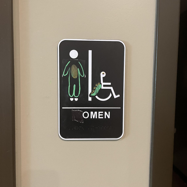 What was formerly a Men's bathroom sign with an icon of a man has been drawn over to make the character genderless and an "O" has been drawn in front of "men" to spell out "Omen."