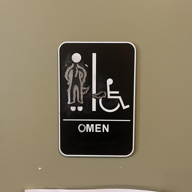 What was formerly a Women's bathroom sign with an icon of a woman has been drawn over to make the character genderless (with a tail) and the "W" in "women" has been covered up so that it reads "Omen."