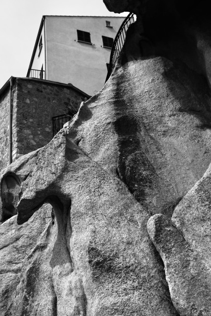 das bild zeigt eine schwarz-weiße nahaufnahme von massiven, zerklüfteten felsen auf korsika. im hintergrund ganz oben im bild ragen die fassaden zweier häuser empor; eines davon besteht aus grobem naturstein, das andere hat eine glatte, helle oberfläche mit dunklen fensterläden. am oberen bildrand verläuft ein schmiedeeisernes geländer entlang der felskante. die struktur des gesteins ist sehr detailreich und zeigt deutliche verwitterungsspuren sowie dunkle streifen, die durch abfließendes wasser entstanden sein könnten. die perspektive ist steil von unten nach oben gerichtet, was die felsen besonders imposant wirken lässt.