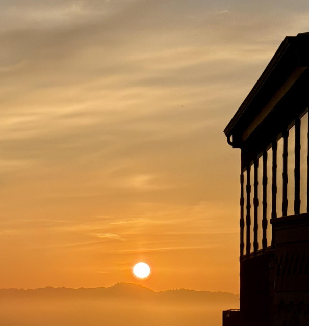A sky just after dawn, and a building at the side with a series of thin vertical windows reflecting it. 

The Sun has just risen above low silhouetted hills, glowing golden orange and surrounded by amber and orange thin hazy clouds. The hills have a layer of amber fog in a line below them, and the sky and wispy clouds above them fade to yellowy gray at the top of the picture.

The building at the side has some angular features, and an eave above the tall windows, making it mostly just a silhouette of geometric shapes. The windows perfectly reflect the amber shades of the sky above the Sun. They are bordered by frames that have small widened elements along the edges in a repeating and complimentary pattern, adding to the geometric feel of the architecture.