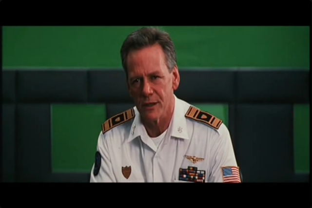 Still from the deleted briefing scene in the film Event Horizon. It shows a late middle-aged grey haired man in a futuristic naval-style uniform with an unprocessed greenscreen set behind him.