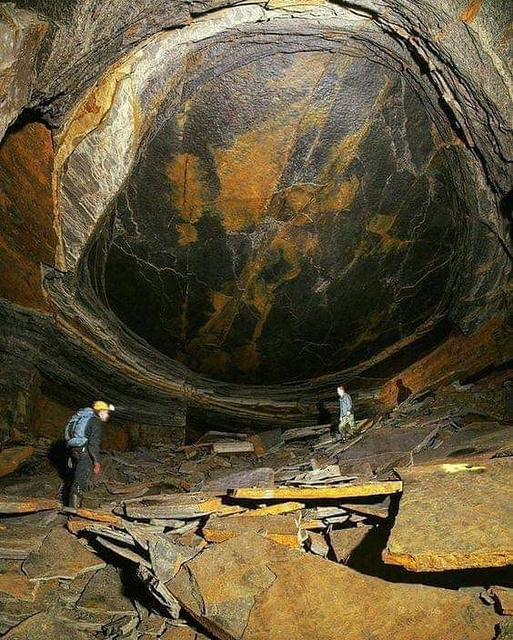A striking photograph taken inside a subterranean stone quarry, looking up at a massive geological formation known as the "Dragon's Eye." The cavern floor is chaotic, cluttered with large, flat, shattered slabs of brown and tan slate and shale rock. Two explorers wearing helmets and headlamps stand among the rubble, their figures providing a dramatic sense of scale. 

Looming directly above them in the curved ceiling of the cavern is a massive, perfectly oval and smooth rock depression that resembles a giant reptilian eye. The "iris" of the eye is textured with rich, vibrant streaks of golden-orange and rust-colored mineral deposits. These bright patterns wrap around a massive, smooth, dark grey and black central dome that forms the "pupil" of the eye. The outer edges of the formation are framed by concentric, layered rings of sediment, making it appear as though a colossal creature is peering down through the rock face.