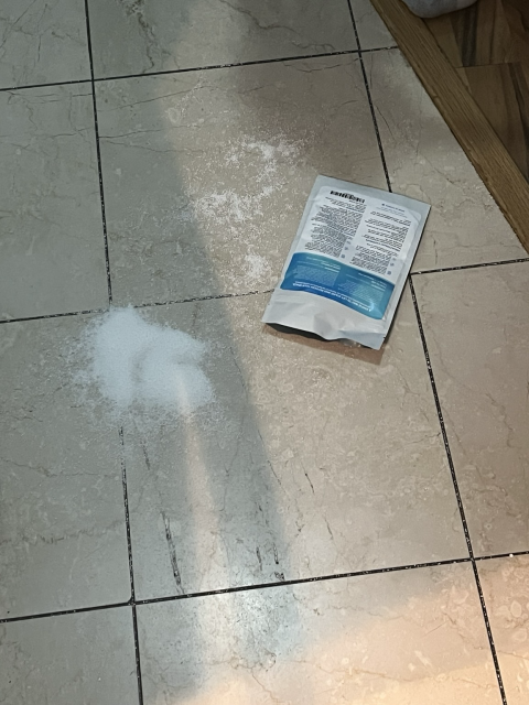 A white and blue bag of sodium percarbonate is on a tiled floor. There’s a pile of white powder beside it. 