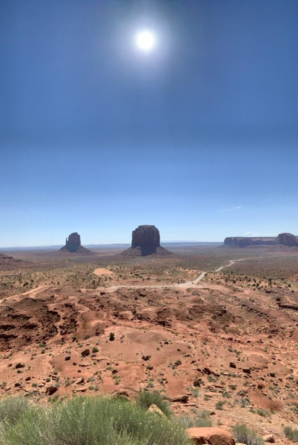 A photo from Monument Valley, Navajo Nation. Looking out over a vast sandstone plateau, several buttes rise dramatically. The sun is centered at the top in a blue sky. The sky contrasts the orange sandstone nicely.