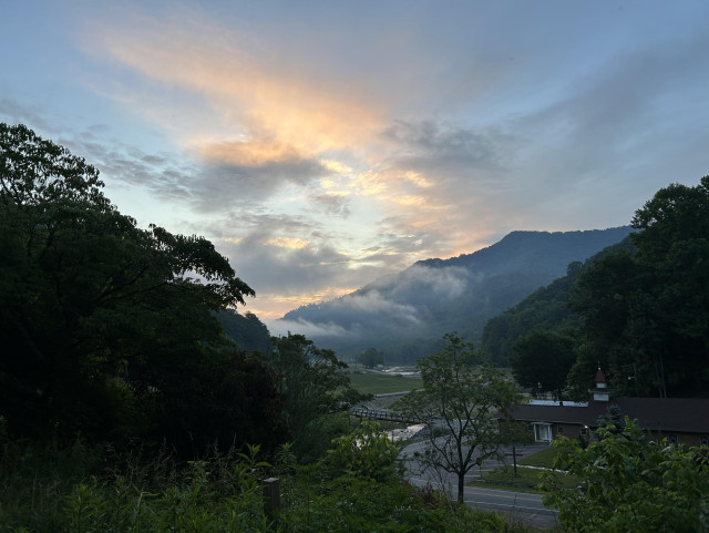 A road meets a river and a chapel sits at the junction as morning breaks with hints of rosy light. Darkest ridge ascends diagonally right to left and is opposed by lighter twin ridges angling upward from center right. Wisps of morning fog laterally punctuate the central blue hued distance and form dynamic clouds that swirl in regular cadence and sweep across the expanse of open sky