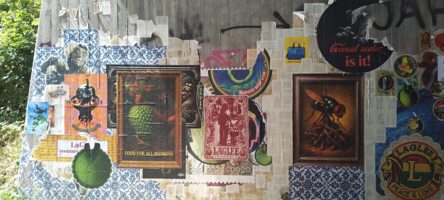 Concrete wall, over and over stuck with paper posters, pages of old books, and other paper decorations. Some paper is eroded or was torn. Others are newer and stuck ontop.

Some of texts read:
"LaGlee nourishing earth"
"Food for all mankind"
"Orchroe not bombs"
"Nature will win"
"LaGlee movements"
"Coconut water is it!"
"Vielfaltigkeit"
"Peace"