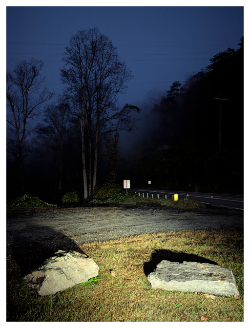 	
A nighttime roadside scene lit by headlights. Two large rocks sit on grassy ground in the foreground. Bare trees rise on the left, fading into mist. A paved road and utility poles curve to the right, disappearing into darkness under a deep blue sky.