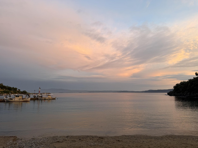 A calm coastal scene at sunset with soft pastel colors in the sky over still water.  
The sky takes up most of the image, filled with broad, streaky clouds lit by the setting sun in shades of peach, pink, and light orange fading into pale blue at the top.  
Below the sky, the sea is very smooth and reflective, tinted with muted pinks and greys from the clouds.  
In the lower part of the image there is a narrow strip of small-pebble and sand beach leading to the water.  
On the left side, a small pier or jetty extends slightly into the water with a few white fishing boats moored alongside it; their reflections are faint on the surface.  
Faint hills of Mount Atos stretch across the horizon in the background, appearing as a dark, hazy line.  
On the far right edge, a rocky, tree-covered shoreline juts into the water, mostly in shadow and forming a dark silhouette against the lighter sky and sea.