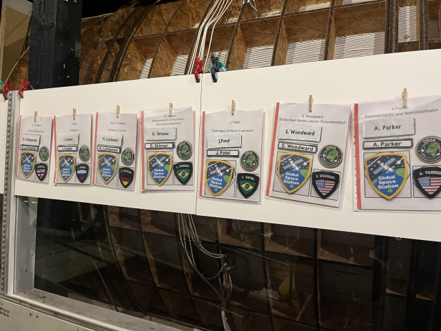 An einem weißen lipbord hängen sieben transparente Umschläge mit Badges und einem Zettel, der einen Namen und einen Beruf angibt. Einer davon ist "Kommandant und Teamstabilisator"
