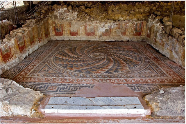 A photograph of an room featuring partially preserved stone walls decorated with faded red,yellow, and white frescoes. The floor is covered with an elaborate mosaic floor with birds and a kantharos, a vessel with two handles, in the centre of a circle of alternating triangles in red, blue, white and ochre, creating an optical illusion of continuous motion, a kind of swirl, surrounded by ornate borders including wave designs. The setting appears to be an archaeological site, with exposed remnants of the original structure visible around the mosaic floor.