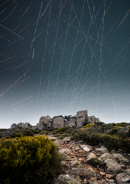 This is closely tied to light pollution: while traditional light pollution comes from ground-based sources, satellites introduce a new, moving form of sky brightness.

AltText:

A striking vertical, long-exposure night photograph capturing an alpine landscape under a sky heavily crisscrossed by satellite trails. In the lower third, a rocky, dirt path winds through low-lying green and golden alpine shrubs toward a prominent outcrop of massive, weathered dolerite boulders. Above this ancient terrain, the dark night sky transitions from a deep teal-blue at the zenith to a hazy, light grey glow along the horizon. The sky is filled with a dense, chaotic grid of straight, intersecting white and faintly colored light trails left by passing satellites. Some trails appear as perfectly spaced dashed lines, slicing across the natural background of faint, static stars and contrasting sharply with the curved, subtle star trails visible on the left edge.