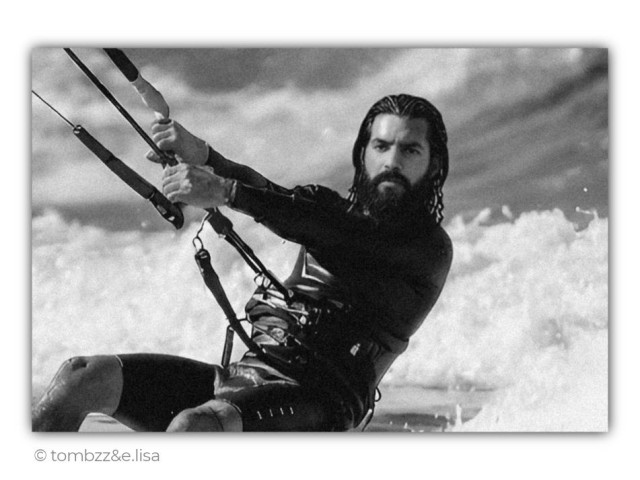 This black-and-white analog photograph shows a dynamic portrait of a kitesurfer at sea. A man in a wetsuit is captured gliding across rough water, gripping the control bar with both hands while the kite lines stretch beyond the frame. His long wet hair and full beard add to the rugged character of the image. His focused gaze is directed toward the horizon, following the wind and waves.
Behind him, the ocean explodes into bright white spray, with foaming waves and airborne droplets creating a strong sense of movement. Heavy clouds fill the sky, reinforcing the atmosphere of open water and untamed natural forces. The contrast between the dark figure and the white surf draws attention to the kitesurfer while preserving the dramatic mood of the scene.
The photograph portrays Tom during a kitesurfing session and was taken by his wife, Elisa.